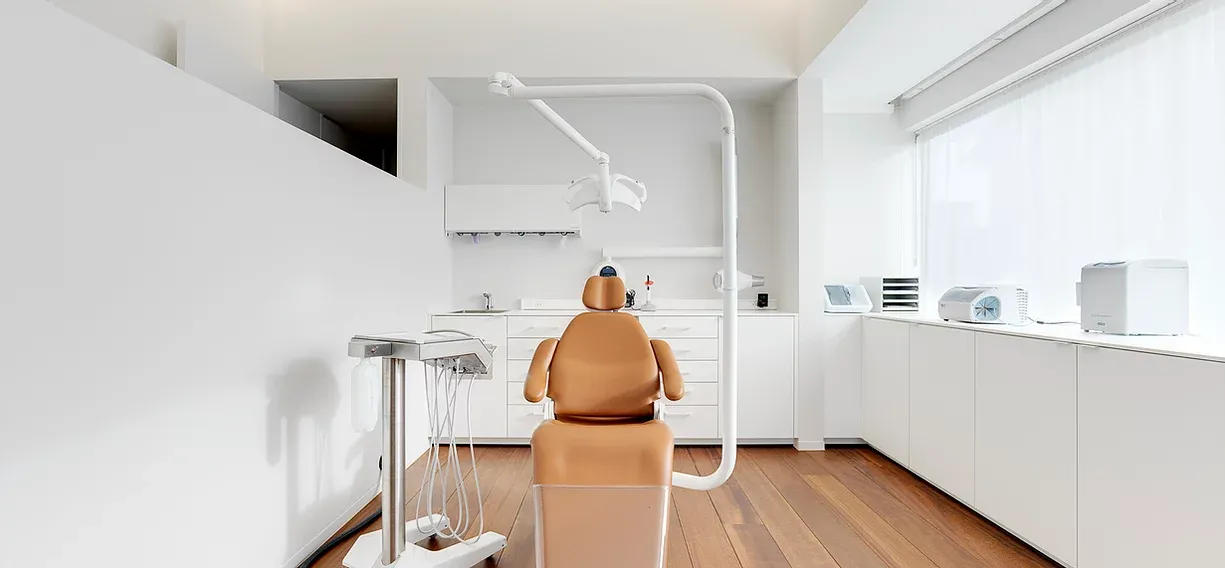 Behandelkamer met wit meubilair, houten vloer en veel lichtinval via grote ramen.